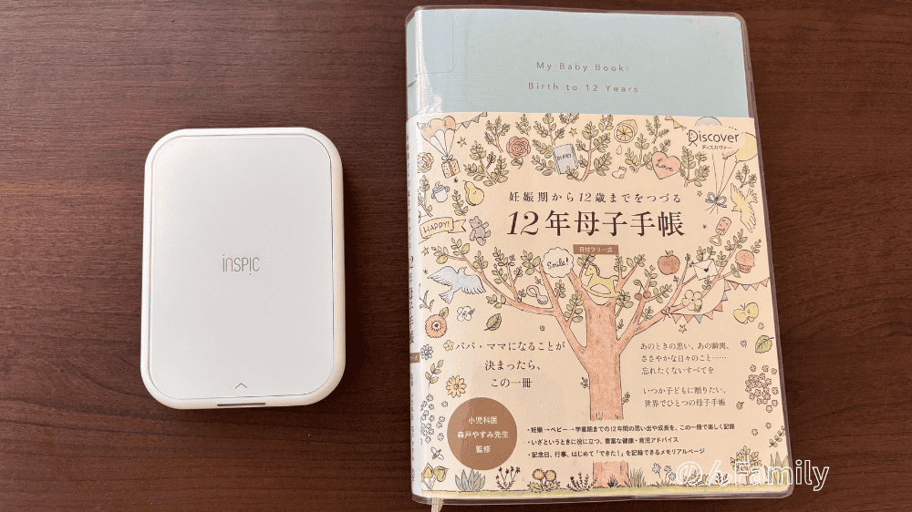 白いiNSPiC PV-223ミニプリンターと、イラスト入りの「12年母子手帳(育児日記)」が並んで置かれている様子。