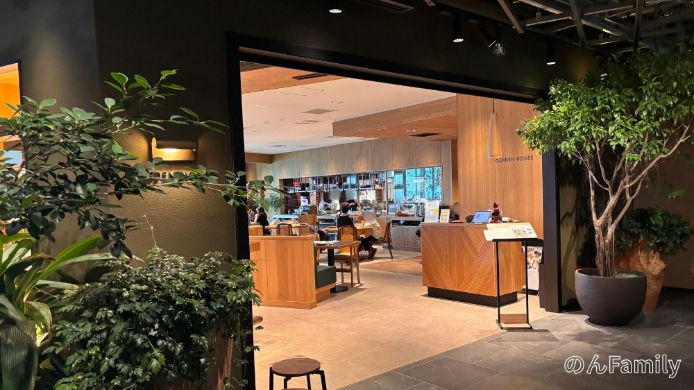 NEWoMan高輪にあるレストラン「GARDEN HOUSE TAKANAWA」の店舗入口。木目調のデザインと、手前に緑豊かな植栽がある。右側には受付カウンターとメニューが置かれ、奥には客席が見える。ベビーカーでも通りやすい広いエントランスの様子。