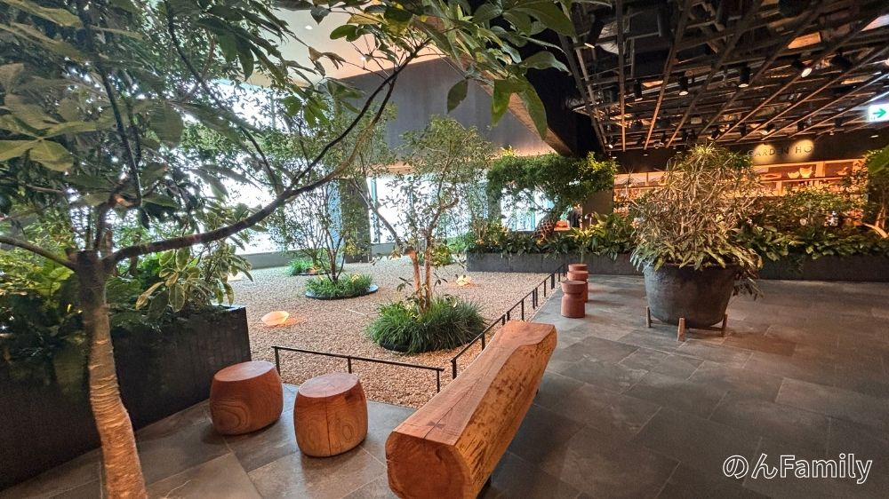 LUFTBAUM高輪の28階にある、緑豊かな屋内庭園の休憩スペース。砂利の敷かれたエリアに、様々な植物が植えられ、その横に丸太のベンチや丸太のスツールが並んでいる。自然を感じられる落ち着いた雰囲気の空間。砂利のエリアを囲む小さなフェンスが見える。