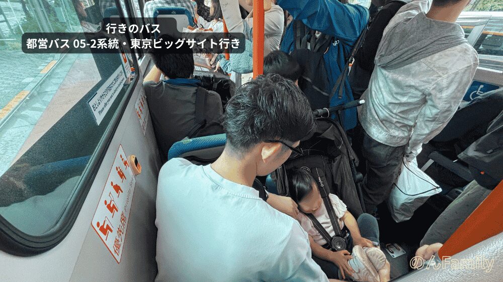 混雑した都営バスの車内で、ベビーカーに乗った1歳児の娘の様子を見守るパパの様子。通路には多くの乗客が立ち並び、車内が満員で混み合っている様子がわかる写真。