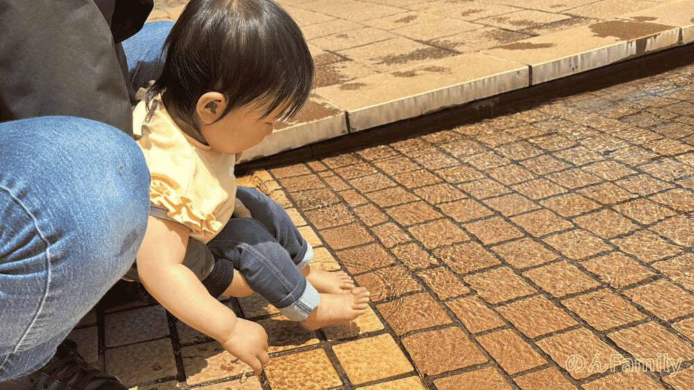 アンデルセン公園のにじの池で、1歳児が池の端に座ってパシャパシャと水遊びを楽しんでいる様子。