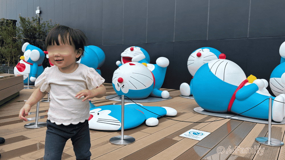 「100％ドラえもん＆FRIENDS（東京ドリームパーク）」の無料エリア、パークB「サイドテラス」。さまざまな寝そべりポーズをした複数のドラえもんフィギュアの前を、元気に歩いて楽しむ1歳児の娘の様子。