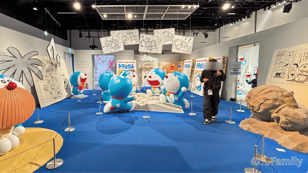 「100％ドラえもん＆FRIENDS」の屋内展示「まんがワールド展」のブース館内。大きなコミックスのオブジェを囲むように配置された複数のドラえもんフィギュアや、壁面に並ぶ藤子・F・不二雄のまんがパネルの様子。
