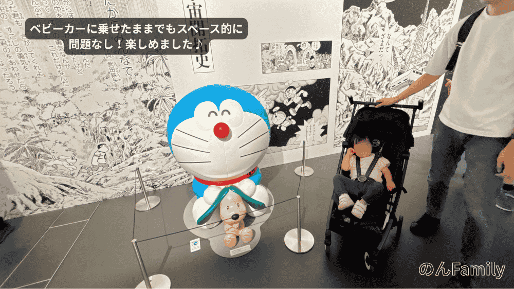 「100％ドラえもん＆FRIENDS」のまんがワールド展の館内で、ベビーカーに乗った1歳児の娘と、ドラえもん・ペコのフィギュア展示を並んで鑑賞する親子の様子。ベビーカーが余裕を持って通れる通路スペースがあることがわかる写真。