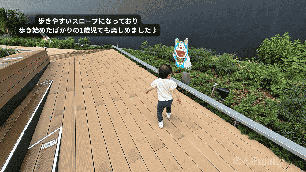 「100％ドラえもん＆FRIENDS」の無料エリア、パークB「サイドテラス」にあるウッドデッキ調の緩やかなスロープ。歩き始めたばかりの1歳児の娘が、のびのびと歩いてお散歩を楽しんでいる後ろ姿の写真。