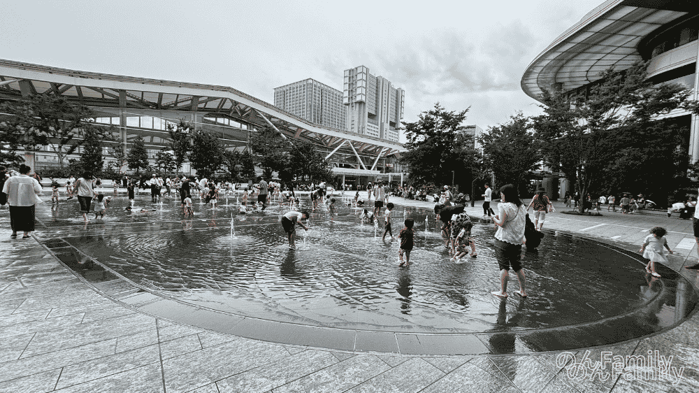 高輪ゲートウェイ駅前の「Gateway Park」で、水遊びを楽しむ子どもたちと見守る親たちの様子。浅い水場で小さな噴水が出ており、夏の子連れ遊び場として賑わっている。