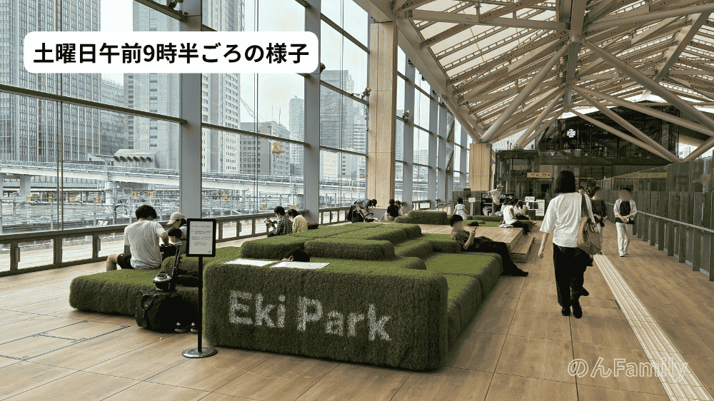土曜日午前9時半の高輪ゲートウェイ駅Eki Park。クッションスペースに大人が座って休憩したり、歩いたりしている。クッションの側面に「Eki Park」の文字。ガラス越しにビル群。画像上部に「土曜日午前9時半ごろの様子」のテキスト。
