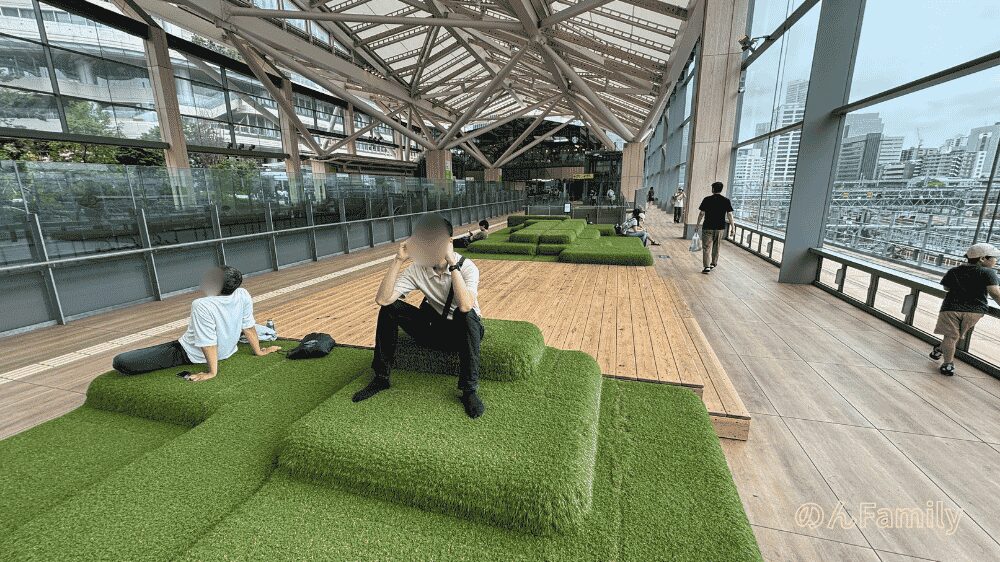高輪ゲートウェイ駅Eki Parkの別アングル。高いトラス構造の天井が印象的。人工芝クッションに座る男性や寝転がる男性。奥に「Eki Park」の文字。ガラス張りの壁面。