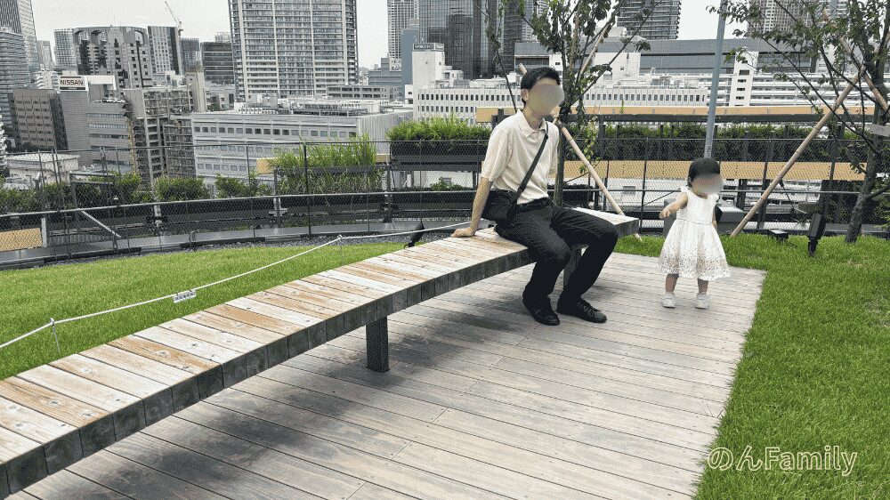 MoN Takanawaのテラスにある木製ベンチ。高輪のビル群を背景に、ベンチに座る大人の隣で1歳くらいの女の子が遊んでいる様子。