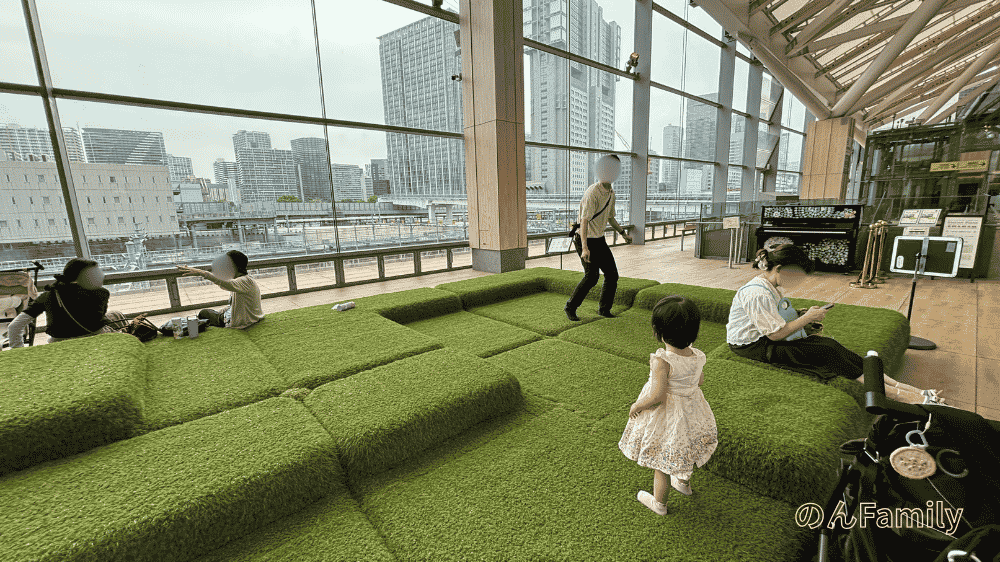 高輪ゲートウェイ駅構内、Eki Park。ガラス張りの広々とした空間で、人工芝のようなクッション遊具で遊ぶ1歳くらいの女の子と見守る大人。背景に線路とビル群。